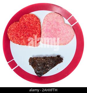 Ein paar herzförmige Plätzchen und ein süsses Stück Schokoladenkuchen Kuchen auf einer weißen Platte in der Form eines Gesichtes für das Feiern Valentines Tag. Stockfoto