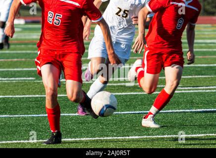 Zwei high school Soccer Spieler in rote Uniformen Steuerung die Kugel auf einem Ausbrechen mit der anderen Mannschaft in weißen Uniformen Jagd nach Ihnen. Stockfoto