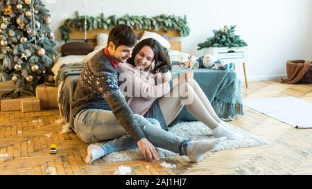 Ein Mann in gemusterten Pullover ist ein Neues Jahr sanft umarmt seine schwangere Frau im Schlafzimmer eingerichtet für das Neue Jahr das Sitzen auf dem Boden neben dem Bett. Mann Stockfoto