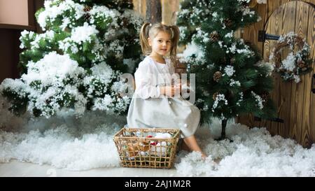 Süße kleine Mädchen in ein leichtes Kleid sitzt auf einem Baumstumpf unter künstlicher Tannen und künstlichen Schnee mit einem Weidenkorb mit Spielzeug. Kinder und Natur Stockfoto