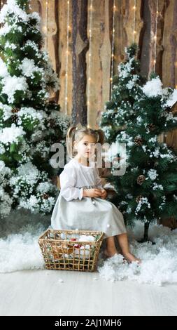 Süße kleine Mädchen in ein leichtes Kleid sitzt auf einem Baumstumpf unter künstlicher Tannen und künstlichen Schnee mit einem Weidenkorb mit Spielzeug. Kinder und Natur. Stockfoto