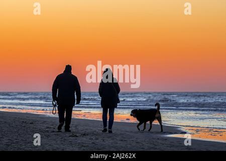 Paar Hundebesitzer zu Fuß entlang der Küste mit unleashed Hund auf Sandstrand, gegen orange Himmel bei Sonnenuntergang Silhouette an einem kalten Abend im Winter Stockfoto