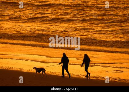 Paar Hundebesitzer zu Fuß entlang der Nordseeküste mit Whippet Hund am Strand, bei Sonnenuntergang Silhouette an einem kalten Abend im Winter Stockfoto