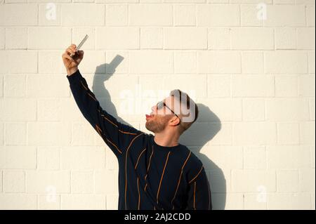 Positive lächelnden Kerl in der casualwear mit Zunge mit Handy unter selfie. Junge bärtige Mann stand im Freien weiße Wand. Männliche selfie, Leis Stockfoto