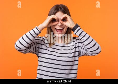 Portrait von Neugierig positive Frau mit braunen Haaren in Long Sleeve Striped Shirt stehend, Hände halten in der Nähe der Augen wie Brillen, Ferngläser Geste. Stockfoto