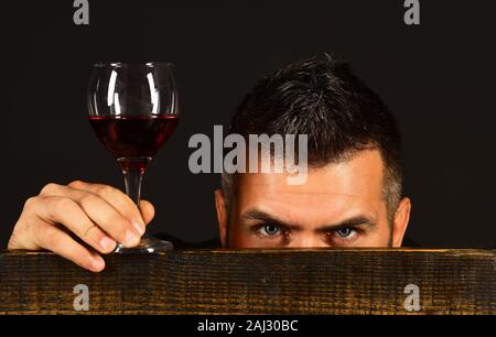 Mann mit Bart hält Glas Wein auf dunklen braunen Hintergrund. Sommelier Geschmack teuer trinken. Degustator mit versteckten Gesicht hinter Holzstuhl. Weinprobe und Verkostung Konzept Stockfoto