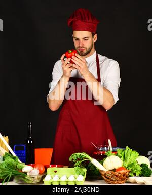 Koch arbeitet in der Küche in der Nähe von Tabelle mit Gemüse und Tools. Koch mit neugierigen Gesicht Red Paprika auf schwarzem Hintergrund hält. Mann in Cook hat und Schürze sieht bei Pepper. Vegetarische Mahlzeit Konzept. Stockfoto