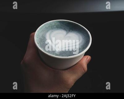 Eine Frau Hand hplding umweltfreundliche Tasse heißen Butterfly pea Latte mit schönen herzförmigen latte Kunst, morgendliche Erfrischung Stockfoto