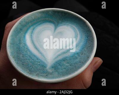 Eine Frau Hand hplding umweltfreundliche Tasse heißen Butterfly pea Latte mit schönen herzförmigen latte Kunst, morgendliche Erfrischung Stockfoto