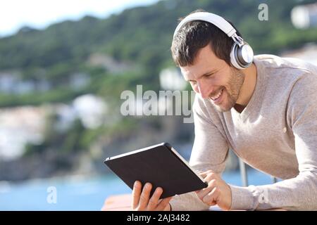 Happy erwachsener Mann, der drahtlose Kopfhörer Kontrolle Tablette Inhalt in einen Balkon am Strand Stockfoto