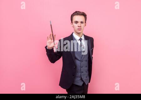 Porträt der jungen Zauberer im Tuxedo stehend mit erhobenen Zauberstab und Kamera mit schweren konzentrierten Ausdruck, Illusionist, Trick. Stockfoto