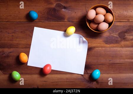 Frohe Ostern Hintergrund, bunte Eier und leeres weißes Papier auf braune Holztisch, Frühling Spaß dekoration design, traditionelle April celebrat Stockfoto