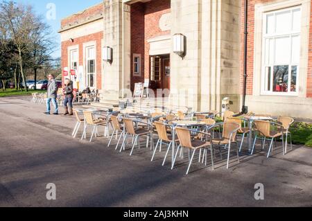 Mann & Frau vorbei gehen. Tische und Stühle draußen ein Cafe an einem sonnigen Wintertag im Stanley Park Blackpool Lancashire England United Kingdom Stockfoto