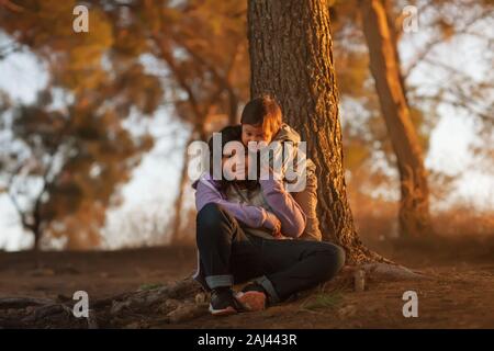 Eine große Schwester und jungen Bruder sitzen in der Nähe von einem Baum auf einem Hügel und umarmen einander, während Sie einen Sonnenuntergang. Stockfoto