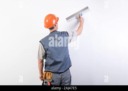 Die Builder glättet die Wand mit einem Spachtel. In Arbeitskleidung und harten Hut. Auf einem hellgrauen Hintergrund. Stockfoto