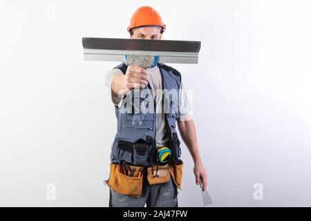 Die Builder verbirgt sein Gesicht hinter einem Spachtel. In Arbeitskleidung und harten Hut. Auf einem hellgrauen Hintergrund. Stockfoto