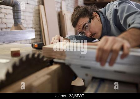 Ernsthafte junge Tischler in schützende Brillen konzentriert sich auf seine Arbeit, die er Arbeiten mit Holz Stockfoto