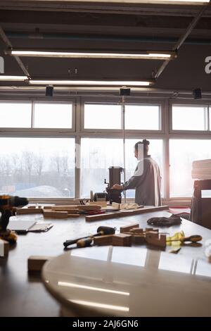 In der Nähe von großer Tisch mit Holzelementen mit Tischler arbeiten an Drehbank vor dem Fenster im Hintergrund Stockfoto