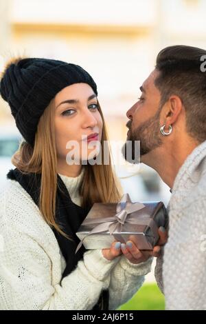Junge Frau in die Kamera schaut gibt einen Silber verpackte Geschenk zu ihrem Freund, während er sie zu küssen. Valentine's Konzept. Stockfoto