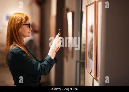 Seitenansicht der jungen Frau, die in der Klage stehen vor der Wand mit Gemälden und Fotografieren der Bilder Stockfoto