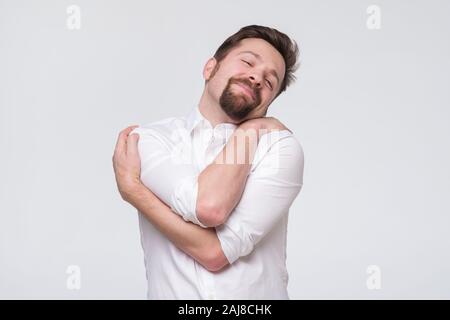 Gerne gut aussehender bärtiger junger Mann sich umarmen, mit geschlossenen Augen und Lächeln genießen. Selbst stolzer Mann Konzept Stockfoto