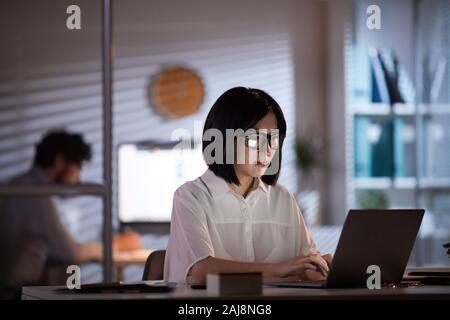 Junge asiatische Frau in Brillen an ihrem Arbeitsplatz sitzen und tippen auf Laptop bis zum Abend im Büro mit ihrer Kollegin im Hintergrund Stockfoto
