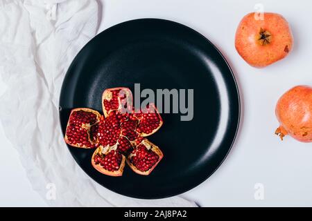 Geschälte Granatapfel auf schwarze Keramikplatte auf weissem Holztisch neben Baumwolltuch und ganze Früchte, Ansicht von oben Stockfoto