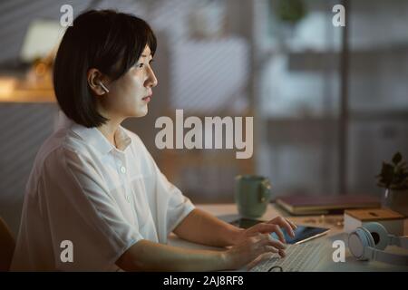 Seitenansicht des Asiatischen junge Frau in drahtlosen Kopfhörern Eingabe über Tastatur am Tisch in dunklen Büro Stockfoto