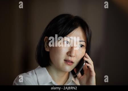 Asiatische junge Frau mit dunklen kurzen Haaren Gespräch am Handy in dunklen Zimmer Stockfoto