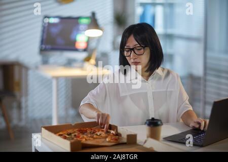 Asiatische junge Geschäftsfrau, ein Stück Pizza, während an Ihrem Arbeitsplatz sitzen und Arbeiten am Laptop. Stockfoto