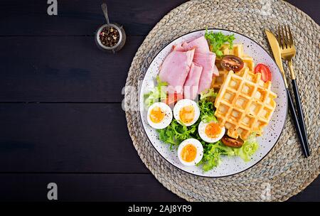 Frühstück mit maismehl Waffeln, gekochtes Ei, Schinken und Tomaten auf dunklem Hintergrund. Vorspeisen, Snack, Brunch. Gesundes Essen. Ansicht von oben, Overhead, Kopieren spac Stockfoto