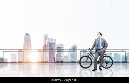 Junger Mann mit Anzug stehen auf Balkon mit dem Fahrrad. Geschäftsmann mit dem Fahrrad auf dem Hintergrund des blauen Himmels über Megalopolis. Männliche Radfahrer halten Stockfoto