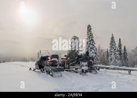 Leute sitzen und auf schneemobilen an der Spitze des Berges sprechen vor dem Hintergrund der Wald und die Sonne im kalten Winter Stockfoto