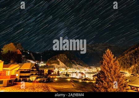 Star Trails über die schneebedeckten Berge und kleinen Dorf Selva di Val Gardena in einer kalten Winternacht Stockfoto