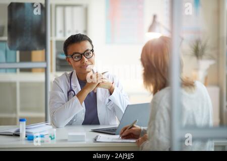 Junge Ärztin im weißen Kittel sitzen am Tisch und Patient sitzt vor ihr im Büro Stockfoto