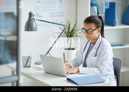 Ernst Ärztin in Brillen und im weißen Mantel sitzen an Ihrem Arbeitsplatz und Tippen auf Laptop im Büro Stockfoto