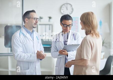 Männlicher Arzt im weißen Kittel im Gespräch mit der reife weibliche Patienten mit Krankenschwester Notizen im Dokument, während Sie im Büro stehen Stockfoto