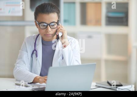 Junge Ärztin in Brillen und im weißen Mantel Gespräch am Handy und suchen bei Monitor des Laptops, während an Ihrem Arbeitsplatz Stockfoto