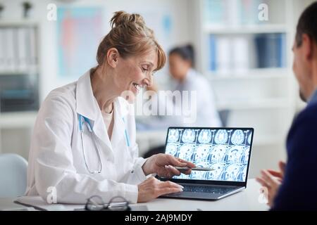 Reife weibliche Arzt sitzen am Tisch und zeigte auf Notebook mit x-ray Bilder und das Gespräch mit dem Patienten im Büro Stockfoto