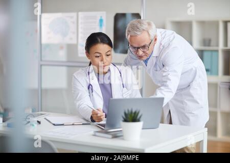 Leitender Arzt im weißen Kittel und junge weibliche Krankenschwester Suche bei Monitor des Laptops und diskutieren online Präsentation im Büro Stockfoto