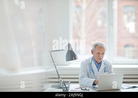 Ernst Senior Medical Specialist am Tisch sitzen und tippen auf Laptop, die er online in seinem Büro arbeiten Stockfoto