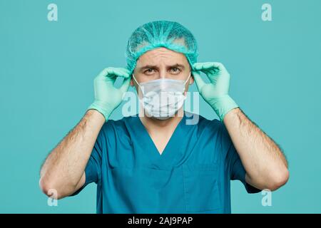 Portrait von Chirurgen in Scrubs und in schützende Maske und Handschuhe in der Kamera, während über dem blauen Hintergrund stehend Stockfoto
