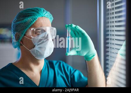 Männlicher Arzt in medizinischen cap Brillen und Schutzmaske holding Reagenzglas in der Hand und Untersuchung von It Stockfoto