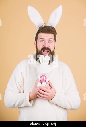 Noch kindisch. Ostern Aktivitäten Konzept. Feiern Sie das Osterfest. Guy bärtigen Hipster fröhliche Osterhase mit langen weißen Ohren. Ostern Kaninchen. Funny Bunny Mann weiche Ohren. Mann mit Kaninchen Anzug spielen mit Spielzeug. Stockfoto