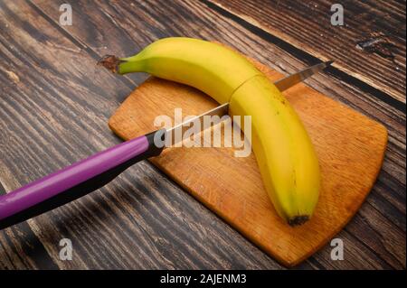Eine reife, gelbe Banane in zwei Hälften schneiden mit einem Messer auf einem Holzbrett auf einer hölzernen Hintergrund. Nahaufnahme Stockfoto