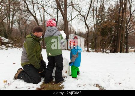 Vater kniet auf dem Boden neben zwei Söhnen, während er einen Schneemann macht Stockfoto