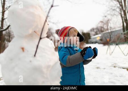 Kleinkind Junge hält Schneeball neben Schneemann, während der Blick nach rechts Stockfoto