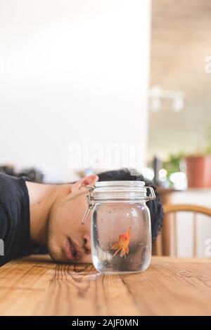 Deprimiert junge liegend auf dem hölzernen Tisch Blick an den Goldfisch im Glas - Einsame Verzweiflung Mann mit seinem Haustier Stockfoto