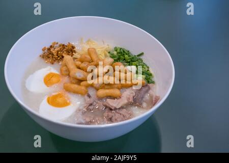 Nahansicht des Reissporridge mit Ingwer, Zwiebeln, gesalzenem Ei, Schweinsrutsche, gebratenem Knoblauch und chinesischen Krückern. Stockfoto