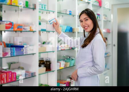 Asiatische junge Frau Apotheker mit einem reizenden Lächeln pick Kapsel Medizin und Kamera in der Apotheke und Drogerie. Medizin, Pharmaindustrie Stockfoto